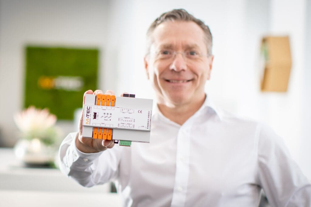 Un hombre sostiene un pequeño aparato. El aparato tiene los colores blanco, naranja, negro y verde. Encima aparece el logotipo de m-tec.