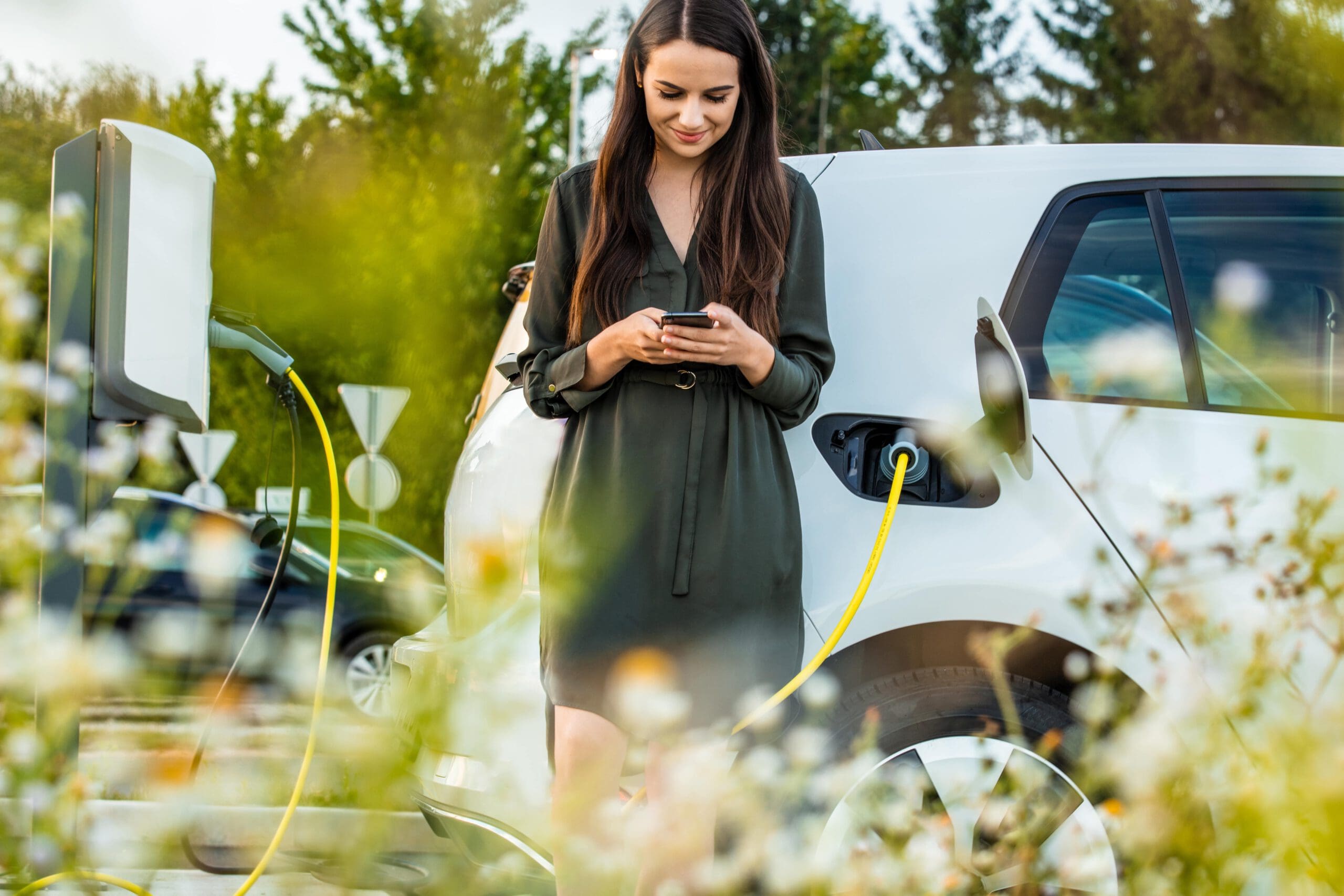 En el centro de la foto aparece una mujer feliz mirando su teléfono. Al fondo, su coche se recarga con un dispositivo blanco.