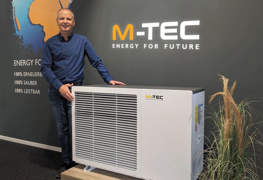 Un hombre está de pie junto a una gran bomba de calor blanca y negra. En el lado derecho de la bomba de calor aparece el logotipo de M-TEC.