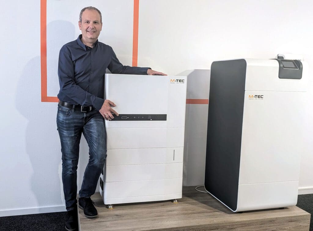Un hombre con camisa de botones azul oscuro y vaqueros está de pie junto a un aparato blanco con un pequeño monitor en la parte frontal de color negro. En la parte superior derecha aparece el logotipo de M-TEC